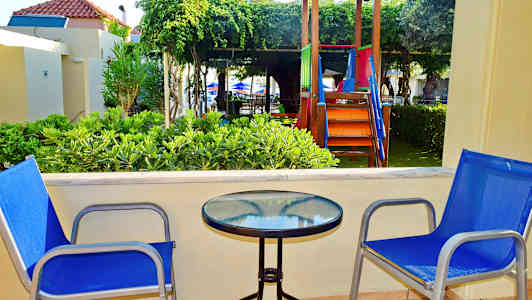 Kaksi sinistä tuolia lasipöydän äärellä terassilla, taustalla leikkipaikka ja vehreyttä. (Tekoälyn luoma teksti)