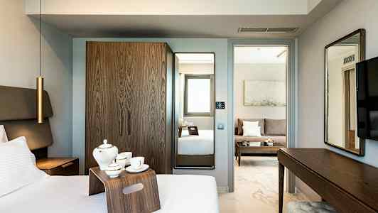 Hotellihuone, jossa on sänky, teetarjotin, vaatekaappi ja peili. Taustalla näkyy istuinalue, jossa on sohva. (Tekoälyn luoma teksti)