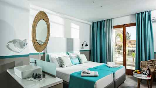 Hotellihuone, jossa on kaksi yhden hengen vuodetta, pesuallas peilin kera ja parvekkeen ovi sinisillä verhoilla. (Tekoälyn luoma teksti)