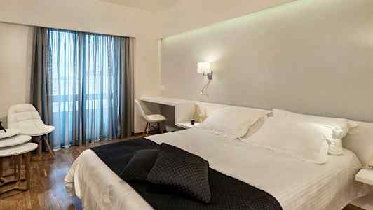 Hotellihuone, jossa on parivuode valkoisilla lakanoilla ja tummilla koristeellisilla tyynyillä. Huoneessa on kirjoituspöytä, valkoiset huonekalut ja lasiovet sinisillä verhoilla. (Tekoälyn luoma teksti)