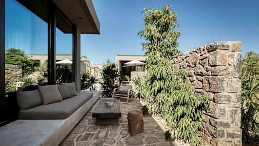 Kivipäällysteinen ulkoterassi, jossa sohva, pöytä ja tuolit lasitalon lähellä, kasvien ja muurien ympäröimänä. (Tekoälyn luoma teksti)