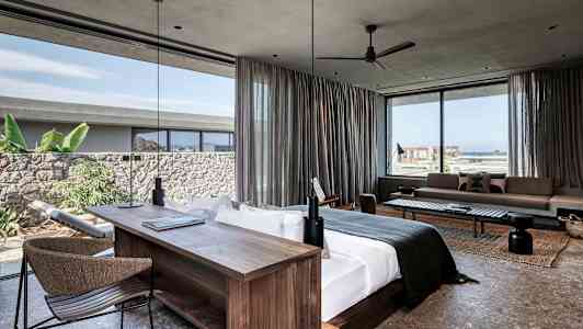 Moderni hotellihuone, jossa suuri sänky, työpöytä ja oleskelualue. Ikkunat, joista näkyy meri ja kasvillisuutta. (Tekoälyn luoma teksti)