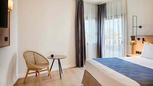 Hotellihuone, jossa moderni tuoli, pieni pöytä ja sänky ikkunoiden vieressä, joissa vaaleat verhot. (Tekoälyn luoma teksti)