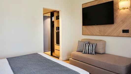 Hotellihuone, jossa on sänky, sohva, seinälle kiinnitetty televisio ja avoin vaatekaappi. (Tekoälyn luoma teksti)