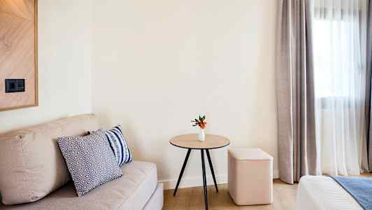 Huone, jossa on beige sohva, puisella pöydällä pieni valkoinen maljakko ja oransseja kukkia, pehmeä jakkara ja verhot ikkunan vieressä. (Tekoälyn luoma teksti)