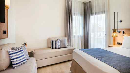 Moderni hotellihuone, jossa on sänky, sohva ja ikkunat vaaleilla verhoilla. (Tekoälyn luoma teksti)