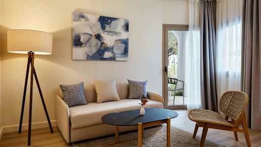 Olohuone, jossa on sohva, pöytä ja tuoli sekä jalkalamppu ja verhot ikkunan edessä. (Tekoälyn luoma teksti)