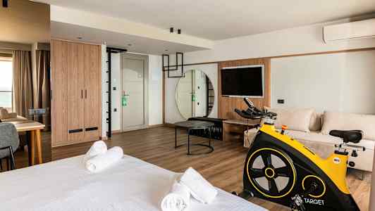 Moderni hotellihuone, jossa on sänky, oleskelualue, kuntopyörä ja televisio. (Tekoälyn luoma teksti)