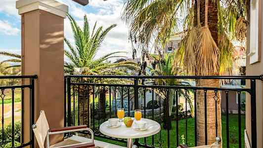 Näkymä parvekkeelta, jossa pöytä, tuolit, juomia ja hedelmiä, sekä taustalla palmuja ja puutarha. (Tekoälyn luoma teksti)