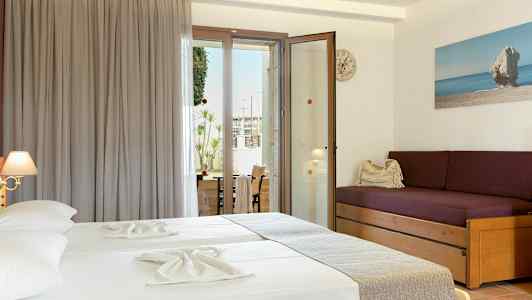 Hotellihuone, jossa kaksi vuodetta vaaleilla lakanoilla, violetti sohva tyynyillä ja lasiovi, joka johtaa parvekkeelle. (Tekoälyn luoma teksti)