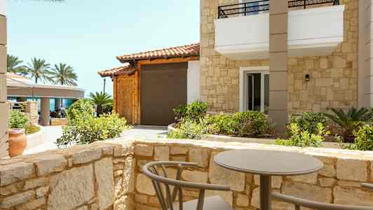 Terassi, jossa on pöytä ja tuolit, joita ympäröi kivimuuri, näkymä rakennukseen, jossa on kivijulkisivu, ja taustalla palmuja. (Tekoälyn luoma teksti)