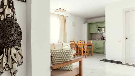 Olohuone, jossa vaalea sisustus, pöytä ja tuolit sekä vihreät keittiökaapit. (Tekoälyn luoma teksti)