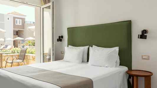 Hotellihuone, jossa sänky ja vihreä sängynpääty, vaaleat seinät ja ikkunasta näkyy terassi. (Tekoälyn luoma teksti)