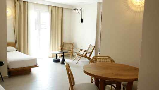 Hotellihuone, jossa on sänky, ruokapöytä ja tuoleja sekä istuinryhmä suuren ikkunan vieressä. (Tekoälyn luoma teksti)