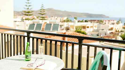 Pöytä, jossa on kirja, lasit ja juoma parvekkeella näkymällä taloihin ja merelle. (Tekoälyn luoma teksti)