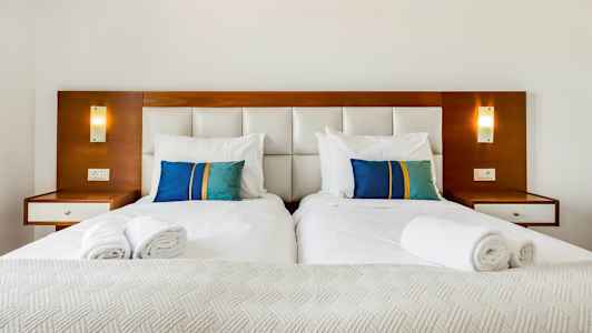 Kaksi erillistä vuodetta valkoisilla vuodevaatteilla ja värikkäillä sinisillä ja vihreillä tyynyillä vuoteen päätyä vasten. (Tekoälyn luoma teksti)