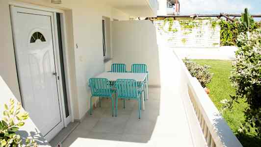 Kuva terassista, jossa on pöytä ja neljä turkoosia tuolia, ympäröimänä vehreyttä. (Tekoälyn luoma teksti)