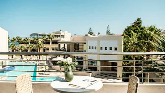 Näkymä parvekkeelta, jossa on pöytä, tuoli ja maljakko kukkineen sekä avoin kirja. Taustalla näkyy rakennus ja aurinkoalue, jossa on uima-allas. (Tekoälyn luoma teksti)