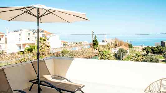 Aurinkotuoleja ja aurinkovarjo terassilla, josta on näkymä merelle ja ympäröiviin rakennuksiin. (Tekoälyn luoma teksti)