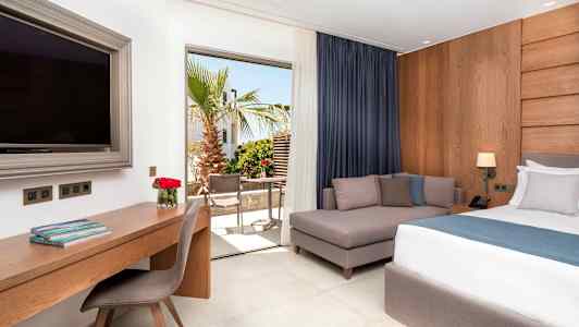 Hotellihuone, jossa on sänky, sohva, työpöytä ja suuri televisio. Näkymä terassille, jossa on puita ja kalusteita. (Tekoälyn luoma teksti)