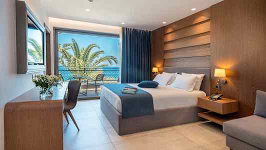 Hotellihuone, jossa on sänky, yöpöytä, työpöytä ja suuret lasiovet, jotka johtavat parvekkeelle palmujen ja meren näkymällä. (Tekoälyn luoma teksti)