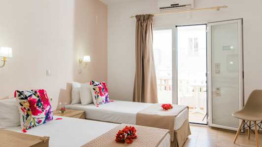 Hotellihuone, jossa on kaksi erillistä vuodetta, koristeltu värikkäillä tyynyillä ja kukilla. Lasinen ovi ja verhot johtavat parvekkeelle. (Tekoälyn luoma teksti)