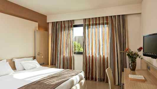 Hotellihuone, jossa on pedattu sänky, kirjoituspöytä tuoleineen, televisio ja ikkuna verhoilla. (Tekoälyn luoma teksti)