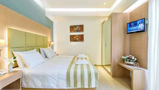 Hotellihuone, jossa on suuri sänky, koristeltu raidallisella päiväpeitolla ja valkoisilla tyynyillä. Seinällä on kukka-aiheista taidetta, ja oikealla näkyy televisio sekä pieni huonekalu, jossa on kukkia. (Tekoälyn luoma teksti)