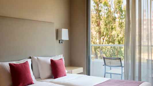 Hotellihuone, jossa on kaksi vuodetta, punaiset tyynyt ja näkymä parvekkeelle, jossa on tuoleja ja puita ulkona. (Tekoälyn luoma teksti)