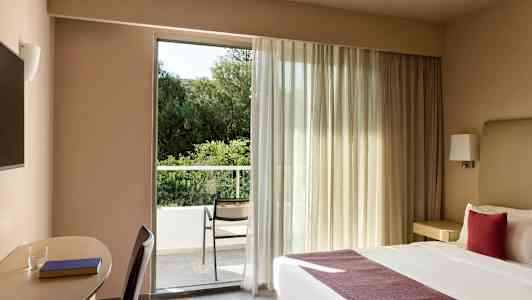 Hotellihuoneessa on parivuode ja työtila sen vieressä. Huoneessa on suuri ikkuna, joka johtaa parvekkeelle, missä on tuoli ja näkymä vehreään maisemaan. (Tekoälyn luoma teksti)