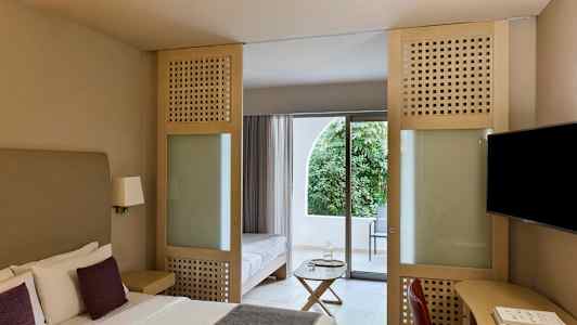 Hotellihuone, jossa on parivuode, istuimet ja lasiovi, joka johtaa parvekkeelle, josta on näkymä vehreyteen. (Tekoälyn luoma teksti)