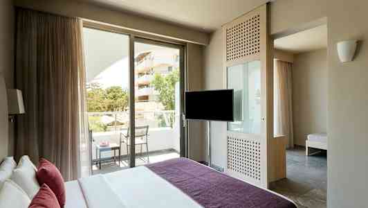 Hotellihuone, jossa on sänky, televisio, parveke tuoleineen ja pöytineen sekä näkymä vihreälle alueelle ja rakennukseen. (Tekoälyn luoma teksti)