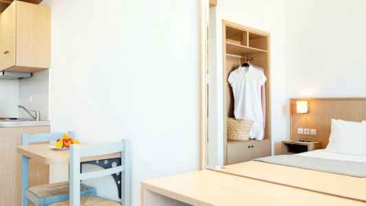 Vaalea hotellihuone, jossa on pieni keittiönurkkaus, pöytä kahdella tuolilla ja sänky. Valkoinen mekko roikkuu vaatekaapissa. (Tekoälyn luoma teksti)