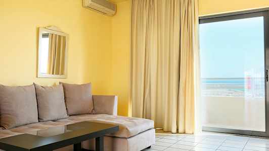 Olohuone, jossa beige kulmasohva, lasipöytä, peili seinällä ja suuret ikkunaverhot näkymällä. (Tekoälyn luoma teksti)