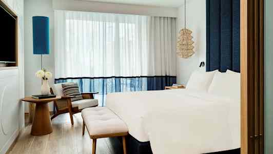 Moderni hotellihuone, jossa on suuri vuode, nojatuoli, pöytä ja kukkia sekä tyylikkäät verhot ja valaistuselementtejä. (Tekoälyn luoma teksti)