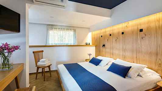 Kuva makuuhuoneesta, jossa on parivuode ja sinisiä yksityiskohtia, puupöytä, tuoli pyyhkeillä ja televisio. (Tekoälyn luoma teksti)