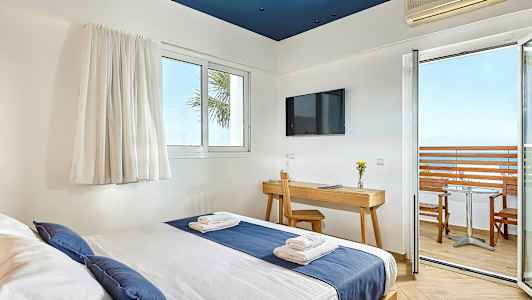 Hotellihuone, jossa sänky sinisen ja valkoisen vuodevaatteet, kirjoituspöytä tuolilla, televisio seinällä ja parveke merinäköalalla. (Tekoälyn luoma teksti)