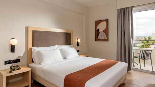 Hotellihuone, jossa on parivuode, valkoiset vuodevaatteet ja ruskea koristelu, yöpöytä puhelimella, seinälamppu sekä taustalla oleva terassi pöydällä ja tuolilla. (Tekoälyn luoma teksti)