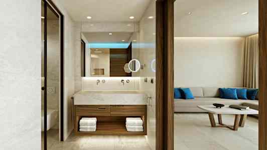 Hotellihuone, jossa on avonainen kylpyhuone, jossa on moderni design, kivipinta ja hyllyt pyyhkeille. Taustalla on vaalea sohvaryhmä, jossa on siniset tyynyt ja sohvapöytä. (Tekoälyn luoma teksti)