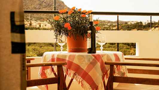 Terassi, jossa pöytä, jossa on ruudullinen pöytäliina, ruukkukasvi, viinipullo ja kaksi lasia. Taustalla kaunis maisema. (Tekoälyn luoma teksti)