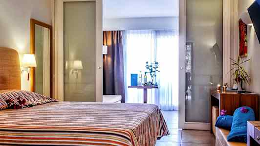 Hotellihuone, jossa on raitapeitteinen sänky. Huoneessa on myös pöytä koriste-esineineen ja taustalla lasiseinät. (Tekoälyn luoma teksti)