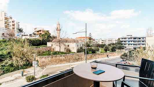 Pöytä, jolla on kahvikuppi ja matkapuhelin, parvekkeella, josta on näkymä vanhaan rakennukseen ja moderneihin taloihin kauempana. (Tekoälyn luoma teksti)