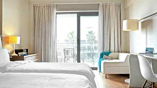 Hotellihuone, jossa valkoiset vuodevaatteet ja sohva lasioven edessä parvekkeelle. (Tekoälyn luoma teksti)