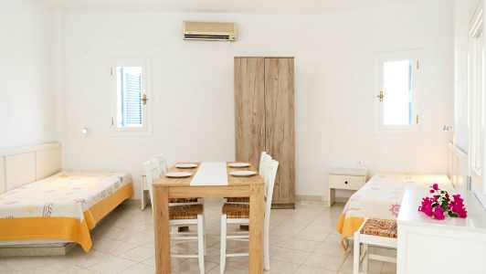 Vaalea huone, jossa on kaksi erillistä vuodetta, ruokapöytä tuoleineen ja vaatekaappi. (Tekoälyn luoma teksti)