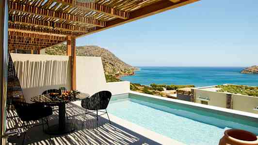 Näkymä terassilta, jossa on uima-allas, pöytä ja tuolit sekä meri ja vuoret taustalla. (Tekoälyn luoma teksti)