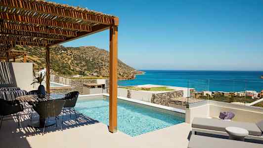 Kuva terassista, jossa on uima-allas, aurinkotuoleja ja ruokailutila, näkymä merelle ja vuorille kirkkaan taivaan alla. (Tekoälyn luoma teksti)