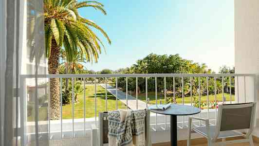 Näkymä parvekkeelta, jolla on pöytä, tuolit ja pyyhe, kohti puutarhaa, jossa on palmuja ja kävelytie. (Tekoälyn luoma teksti)