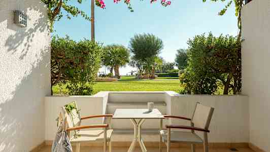 Terassi, jossa kaksi tuolia ja pöytä, joka liittyy vihreään puutarhaan, taustalla palmuja. (Tekoälyn luoma teksti)