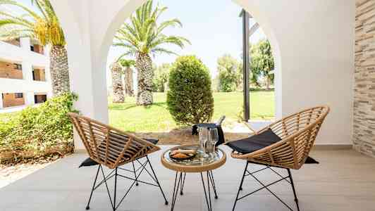 Kaksi punottua tuolia ja pöytä, jossa on lasi ja lautanen terassilla, näkymä vihreään puutarhaan, jossa palmuja. (Tekoälyn luoma teksti)