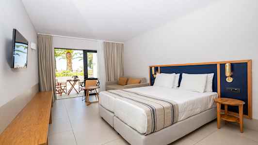 Hotellihuone, jossa iso sänky, istuinryhmä ja näkymä terassille ja puutarhaan. (Tekoälyn luoma teksti)